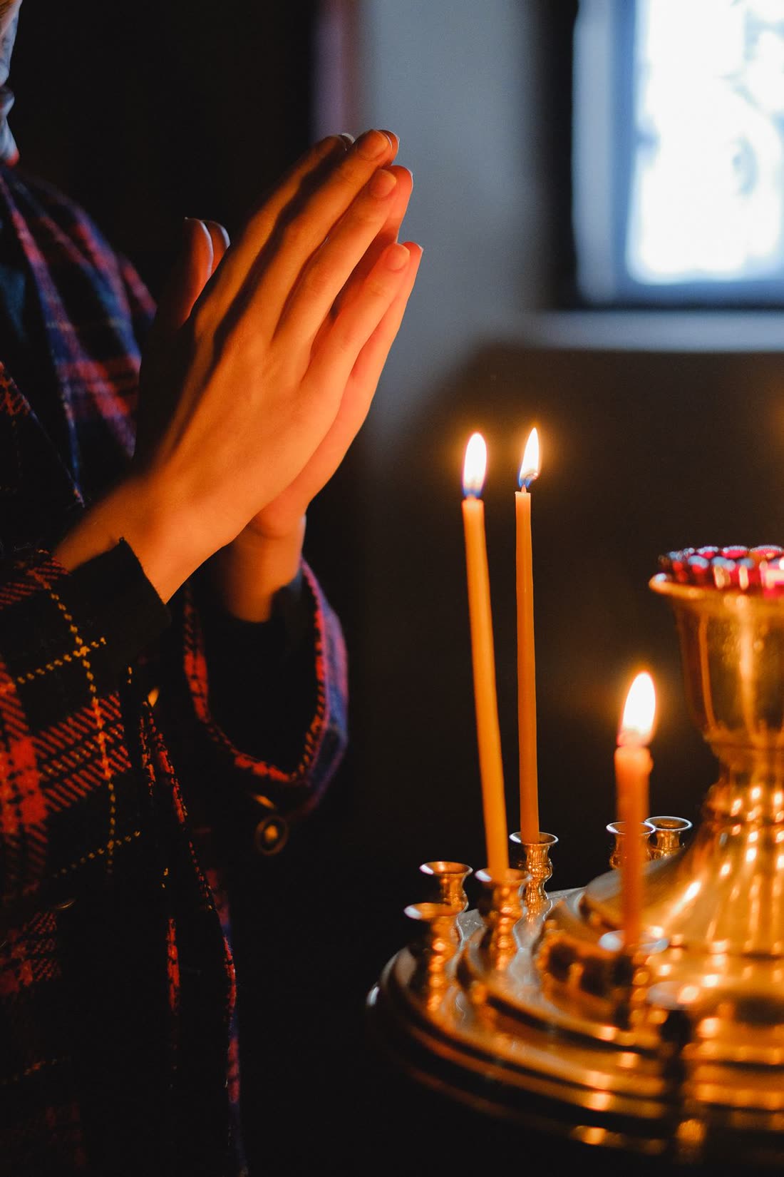 Mãos unidas em oração diante de velas acesas, em luz quente e ambiente escuro
